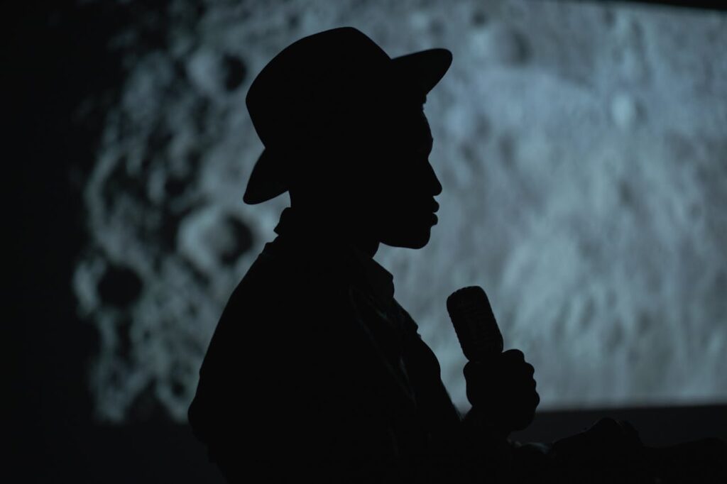 pexels photo 7611745 A dramatic silhouette of a person with a hat holding a microphone against a moon-like background.